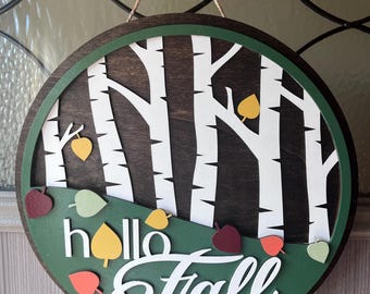 Könnte beinhalten: Ein runder Holztürhänger mit grünem Hintergrund und den Worten "hello fall" in Weiß. Der Hintergrund zeigt eine dunkelbraune Holzmaserung mit weißen Birken und bunten Herbstblättern.