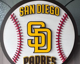 Könnte beinhalten: Runder Holzschild mit dem Logo und Text der San Diego Padres. Das Schild hat ein Baseball-Design mit roten Nähten, einem weißen Hintergrund und braunen und gelben Buchstaben. Ein Seil ist zum Aufhängen befestigt.