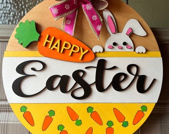 Könnte beinhalten: Rundes Ostern-Holzschild mit einer rosa und Jute-Schleife. Es zeigt eine Karotte mit dem Wort "HAPPY" und einem Hasen. Das Wort "EASTER" ist darunter abgebildet, mit Karotten am unteren Rand.
