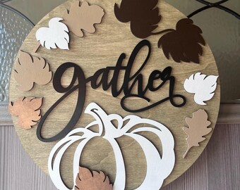 Könnte beinhalten: Runder Holzschild mit dem Wort "gather" in schwarzer Schrift, einem weißen Kürbis und Blattausschnitten in Braun- und Weißtönen. Für Herbstdekoration.