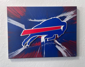 Könnte beinhalten: Leinwandkunst mit dem Logo der Buffalo Bills. Das Logo ist ein blauer Büffel mit einem roten Streifen, vor einem blauen Hintergrund mit roten und weißen Pinselstrich-Akzenten. Das Kunstwerk ist auf einer rechteckigen Leinwand montiert.