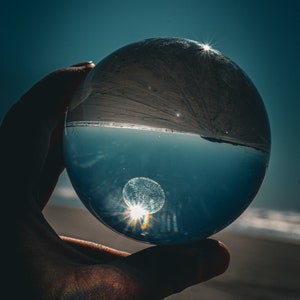 Puede incluir: Una esfera de vidrio transparente sostenida en una mano, que refleja una escena de playa con un sol brillante. La esfera distorsiona la vista, creando una perspectiva única del cielo, la arena y el agua. La imagen tiene una calidad cinematográfica y artística.