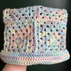 May include: A crocheted cowl in shades of blue, green, and pink. The cowl is made with a granny square stitch and has a simple border.