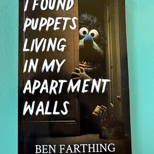 May include: Book cover with a black background and white text that reads "I found puppets living in my apartment walls" and "Ben Farthing". The cover features a photo of a blue and brown puppet peeking out from behind a door.