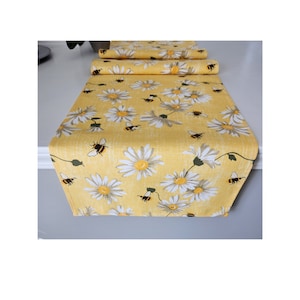 May include: A yellow table runner with a repeating pattern of white daisies and black and yellow bees. The runner is made of fabric and is folded over on itself in the image.