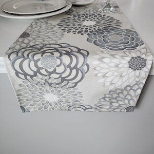 May include: A beige table runner with a floral pattern in shades of gray. The flowers are large and have a layered, textured look.