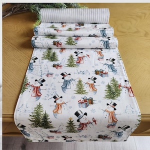 May include: A white table runner featuring a repeating pattern of snowmen, pine trees, and presents. The snowmen are wearing black top hats and scarves. The background is a light blue with snowflakes.