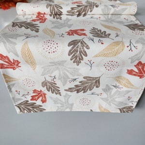 May include: A white table runner with a fall leaf pattern. The leaves are in shades of brown, red, and gold. There are also small red berries and white dots on the runner.