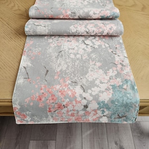 May include: A gray table runner with a floral pattern featuring pink and blue flowers. The runner is made of a soft, textured fabric.