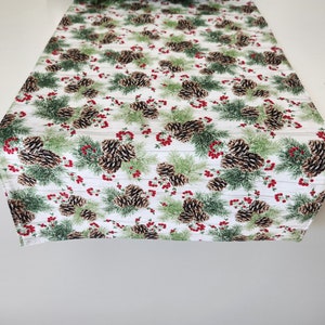 May include: White table runner with a repeating pattern of pine cones, pine branches, and red berries.