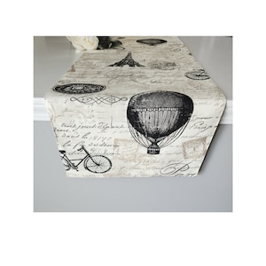 May include: A black and white table runner with a vintage Parisian theme. The runner features a hot air balloon, a bicycle, and the Eiffel Tower. The background is a cream colour with black script writing.
