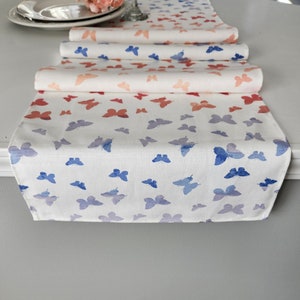 May include: A white table runner with a repeating pattern of blue butterflies. The butterflies are faded in a watercolor style, creating a gradient effect. The runner is folded in thirds, with the top layer showing a lighter shade of blue.