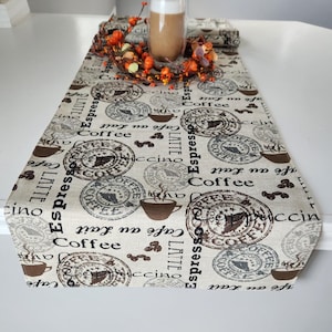 May include: A beige table runner with a coffee theme. The runner features a repeating pattern of coffee cups, coffee beans, and the words "Espresso", "Coffee", "Latte", and "Cappuccino".