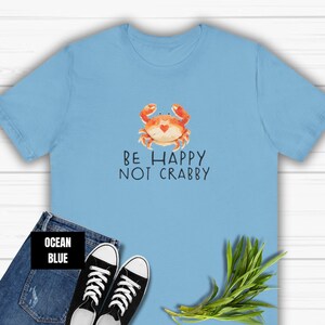 May include: A light blue t-shirt with a cartoon crab and the text "Be Happy Not Crabby" printed on the front. The shirt is displayed on a wooden surface with a pair of blue jeans and a green plant.