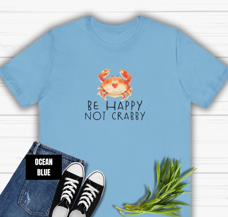 May include: A light blue t-shirt with a cartoon crab and the text "Be Happy Not Crabby" printed on the front. The shirt is displayed on a wooden surface with a pair of blue jeans and a green plant.