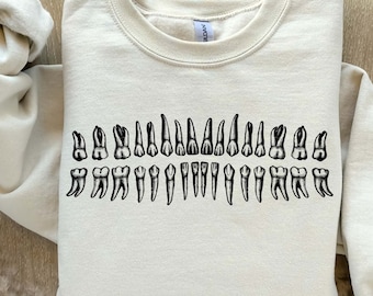 Peut inclure: Un sweat-shirt beige avec un motif noir représentant une dentition humaine complète. Les dents sont disposées en rangée, montrant les incisives, les canines, les prémolaires et les molaires.