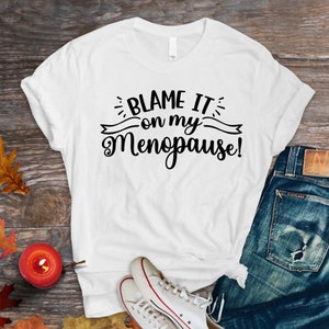 May include: White t-shirt with the text "BLAME IT on my Menopause!" in black script. The shirt is paired with blue jeans and white sneakers with red accents. A small candle and autumn leaves are also visible.