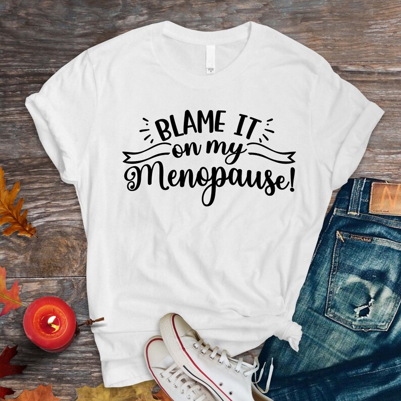 May include: White t-shirt with the text "BLAME IT on my Menopause!" in black script. The shirt is paired with blue jeans and white sneakers with red accents. A small candle and autumn leaves are also visible.