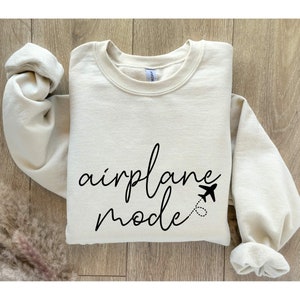 Puede incluir: Una sudadera de color crema con el texto "airplane mode" en letras cursivas negras. Un peque&ntilde;o icono de avi&oacute;n est&aacute; al lado del texto.