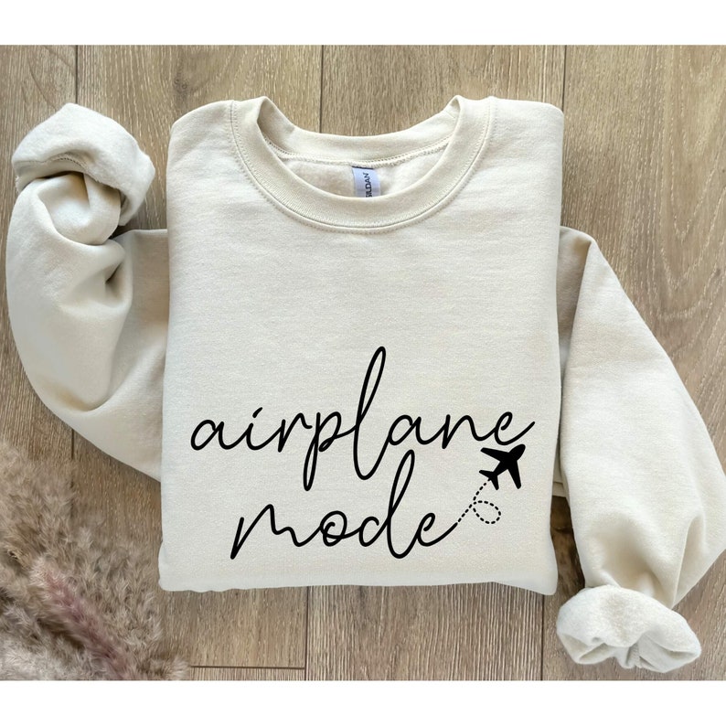Puede incluir: Una sudadera de color crema con el texto "airplane mode" en letras cursivas negras. Un peque&ntilde;o icono de avi&oacute;n est&aacute; al lado del texto.