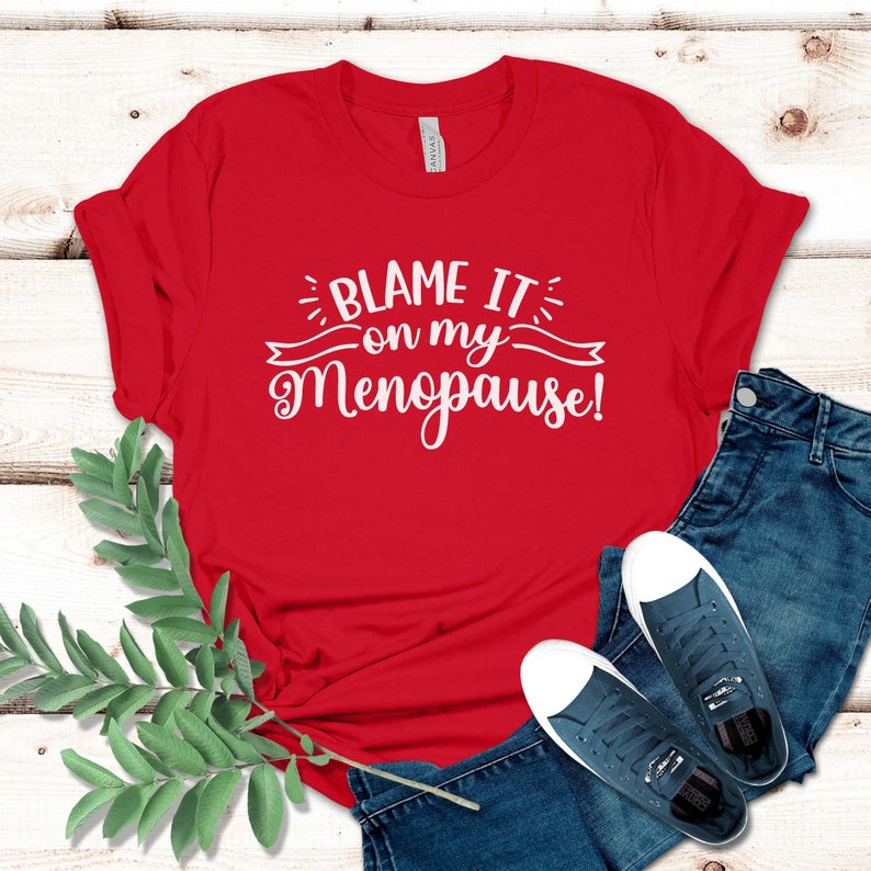 May include: A red t-shirt with the text "Blame it on my Menopause!" in white script. The shirt is paired with blue jeans and navy blue sneakers with white soles. A sprig of green leaves is in the lower left corner.