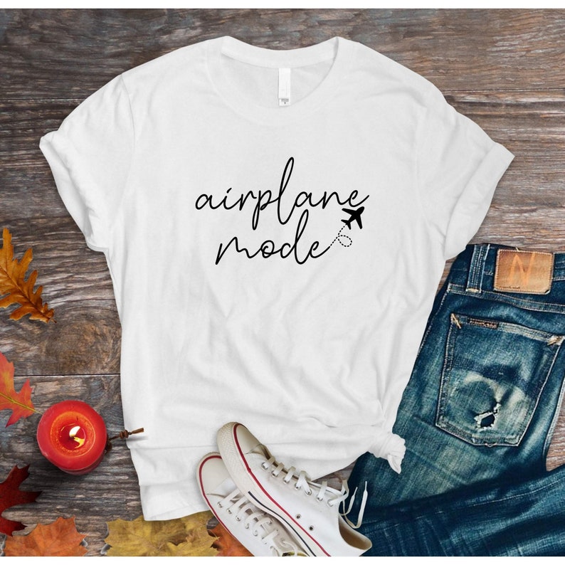 Puede incluir: Camiseta blanca con el texto "airplane mode" en escritura cursiva negra. Un peque&ntilde;o icono de avi&oacute;n est&aacute; al lado del texto.