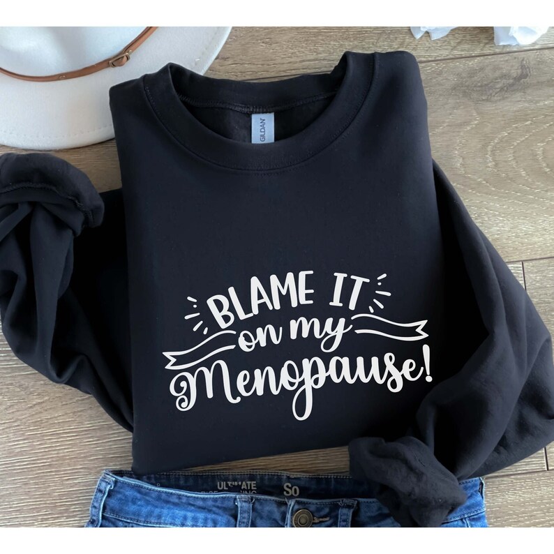 May include: Black sweatshirt with the phrase "BLAME IT on my Menopause!" in white script. The sweatshirt is folded, with a pair of blue jeans visible at the bottom. A white hat is visible in the background.