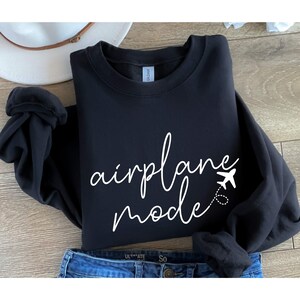 Puede incluir: Sudadera negra con texto blanco que dice "modo avi&oacute;n" y un peque&ntilde;o gr&aacute;fico de avi&oacute;n.