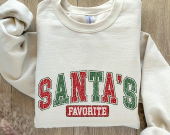 Peut inclure: Sweat-shirt crème avec l'inscription "SANTA'S FAVORITE" en lettres rouges et vertes. Le sweat-shirt est plié, mettant en valeur le motif de vacances festif. Le motif est centré sur le devant du vêtement.