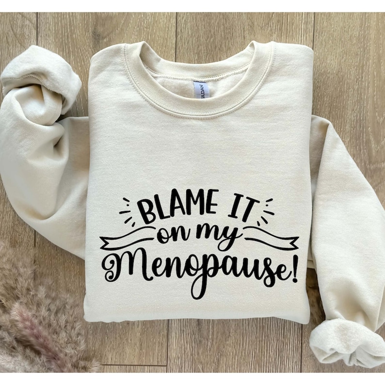 May include: A cream-colored sweatshirt with the phrase "BLAME IT on my Menopause!" in black script. The text is centered on the front of the garment. The sweatshirt is folded, with the sleeves visible.