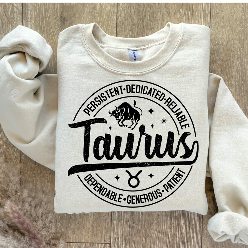 May include: A cream-colored sweatshirt with a black circle graphic featuring the word "Taurus" in a vintage font. The circle also includes the Taurus zodiac symbol and the words "Persistent, Dedicated, Reliable, Dependable, Generous, Patient".