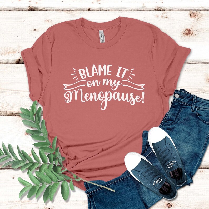 May include: A terracotta-colored t-shirt with the text "BLAME IT on my Menopause!" in white script. The shirt is paired with blue jeans and navy blue sneakers with white toes. A sprig of green leaves is in the lower left corner.