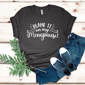 May include: Dark gray t-shirt with the text "BLAME IT on my Menopause!" in white script. The shirt is paired with blue jeans and navy blue sneakers with white soles. A sprig of green leaves is on the left.