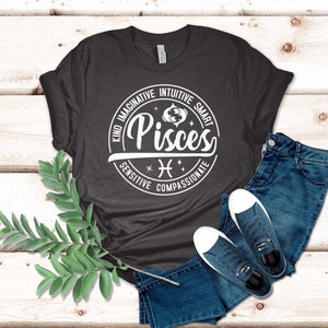 Może przedstawiać: Szary t-shirt z białym kołem z napisem "Ryby" z symbolem zodiaku Ryb i słowami "Miły, Wyobraźnia, Intuicja, Inteligentny, Wrażliwy, Wsp&oacute;łczujący".