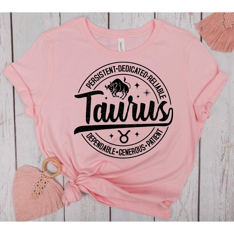 May include: Pink t-shirt with a black circle graphic featuring the zodiac sign Taurus. The circle includes the words "Taurus", "Persistent", "Dedicated", "Reliable", "Dependable", "Generous", and "Patient".