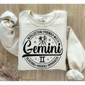 May include: A cream-colored sweatshirt with a black circle graphic featuring the Gemini zodiac sign and the text "Gemini II". The circle also includes the words "Intellectual, Friendly, Witty, Adjustable, Versatile, Enthusiastic".