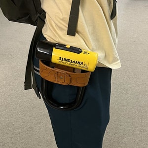 Puede incluir: Una persona que lleva una camiseta amarilla con el texto "Music & Respect" y una mochila verde, con un candado en U Kryptonite amarillo y negro sujeto a su cinturón.