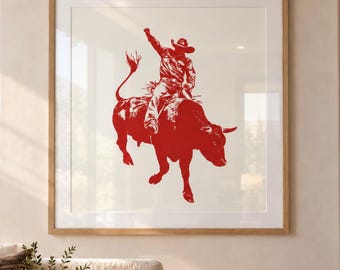 Può includere: Un'opera d'arte da parete quadrata incorniciata che presenta una grafica rossa di un cowboy che cavalca un toro imbizzarrito. L'immagine è centrata all'interno di un passe-partout chiaro e di una semplice cornice in legno, con il cavaliere.