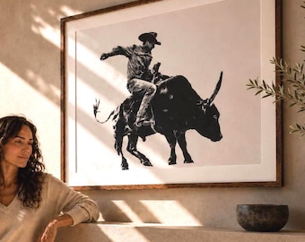 May include: A framed black and white print depicts a cowboy riding a bull. The artwork is in a dark wooden frame and hangs on a neutral-colored wall. A woman is seen in the foreground, leaning on a ledge. A small, dark bowl sits on the ledge.