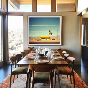 May include: A modern dining room with a wooden table and chairs. The table is set with placemats, plates, and silverware. A large framed photograph of a yellow building in a desert landscape hangs on the wall.