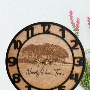 May include: A wooden clock with black numbers and a black frame. The clock face has a woodgrain background with a laser-etched image of trees and the text "Nearly Home Trees".