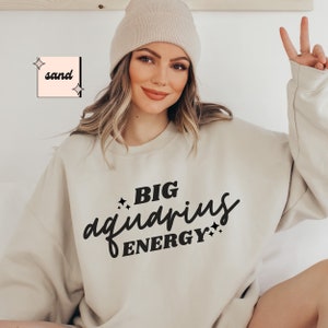 May include: A beige sweatshirt with the text "BIG aquarius ENERGY" in black lettering. The sweatshirt has a relaxed fit and is worn by a person with long brown hair and a beige beanie.