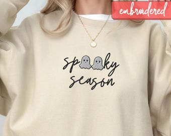 May include: A beige sweatshirt with the words "spooky season" embroidered in black script, featuring two ghost figures. The word "embroidered" is in a red rectangle in the upper right corner. A gold necklace is visible.