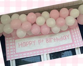 May include: Hand-painted pink and white gingham birthday banner with the text "Happy 1st Birthday". The banner is decorated with a garland of pink and white balloons.