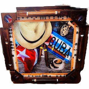 May include: A wooden-framed display featuring Cuban-themed items. A straw hat, cigars, a Cuban flag, and a sign reading "CUBA" are arranged on a wooden surface. A cup of coffee and a decorative percussion instrument are also present.