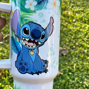 Peut inclure: Un gobelet en acier inoxydable blanc avec un personnage bleu Stitch de Lilo & Stitch de Disney. Le gobelet a un motif floral tropical avec des feuilles vertes et des fleurs blanches.