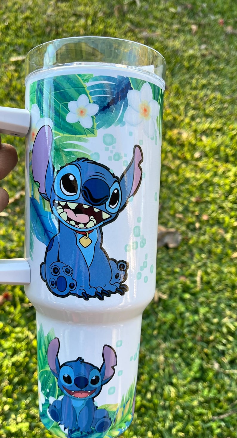 Peut inclure: Un gobelet en acier inoxydable blanc avec un personnage bleu Stitch de Lilo & Stitch de Disney. Le gobelet a un motif floral tropical avec des feuilles vertes et des fleurs blanches.