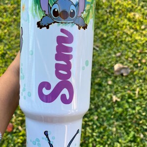 Peut inclure: Un gobelet en acier inoxydable blanc avec un personnage bleu Stitch de Lilo & Stitch. Le gobelet a un motif de feuilles tropicales vertes et blanches et le nom "Sam" &eacute;crit en violet.