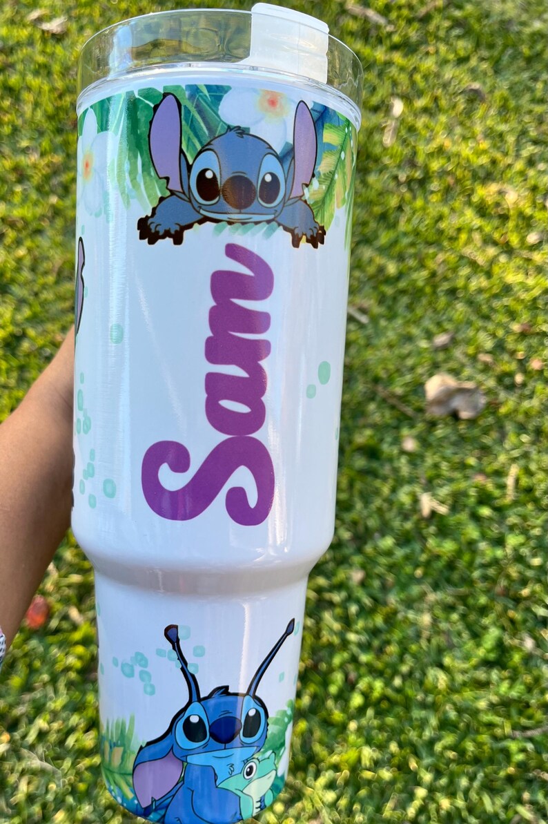 Peut inclure: Un gobelet en acier inoxydable blanc avec un personnage bleu Stitch de Lilo & Stitch. Le gobelet a un motif de feuilles tropicales vertes et blanches et le nom "Sam" &eacute;crit en violet.