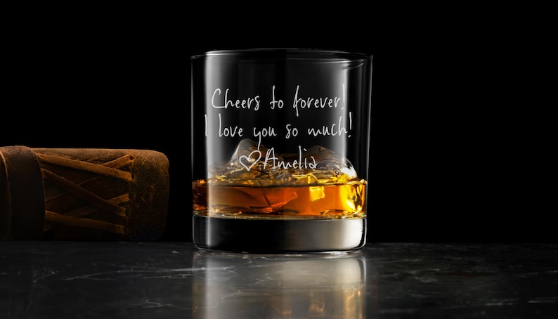 May include: A clear glass tumbler filled with amber liquid and ice cubes, featuring white engraved text that reads: Cheers to forever! I love you so much! with a heart symbol and the name Amelia written in a cursive script style font.